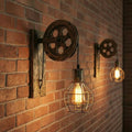 Industrial-style wall sconces mounted on a red brick wall, featuring dark wood wheel hardware and hanging Edison-style bulbs in wire cages, with warm light glowing against the wall.