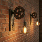 Industrial-style wall sconces mounted on a red brick wall, featuring dark wood wheel hardware and hanging Edison-style bulbs in wire cages, with warm light glowing against the wall.