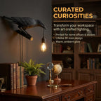 A black raven lamp with a lit bulb hangs in a home office with books and a globe on a wooden desk.