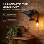 A black raven lamp with a lit bulb is mounted on a wooden wall above a plant and books.