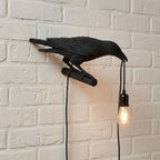 A black raven lamp with a lit bulb is mounted on a white brick wall.