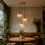 Three-light pendant ceiling fixture with ribbed clear glass shades and warm brass-toned caps, hanging over a round wooden dining table in a cozy interior setting.