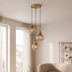 A modern three-light pendant ceiling fixture with ribbed amber-tinted glass shades and a brushed brass finish, hanging in a bright, contemporary living room.