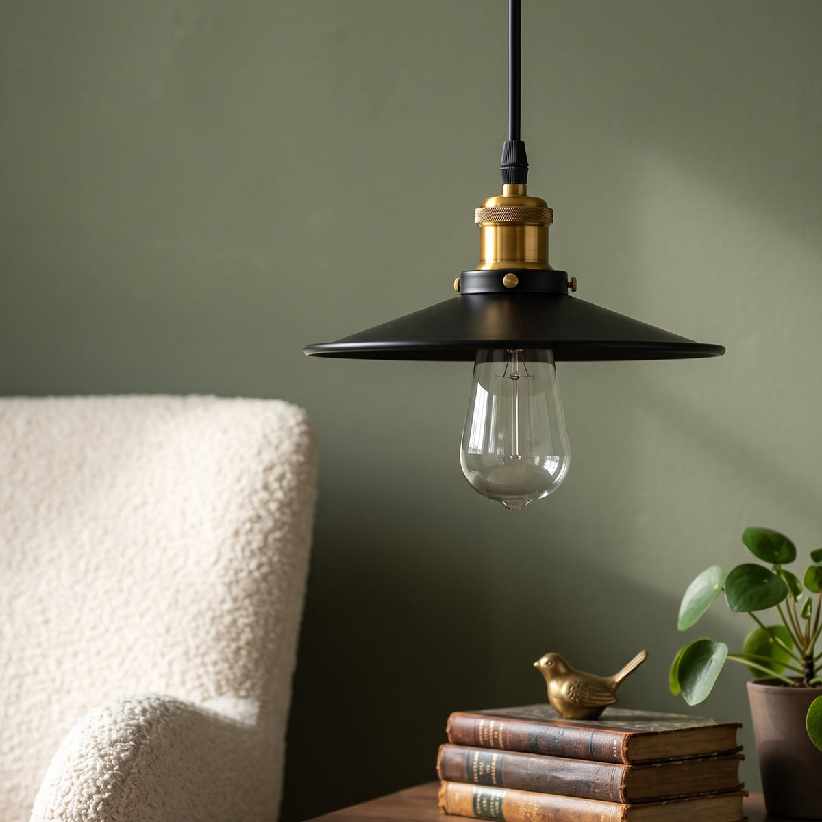 A black metal pendant light with a brass-finished socket and a clear glass bulb, hanging in a styled room setting next to a cream boucle chair, books, and a small plant.