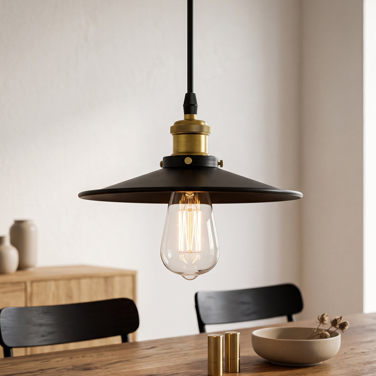 A black metal pendant light with a wide conical shade, a brass-colored socket, and a clear glass bulb hanging over a wooden dining table in a softly lit interior.