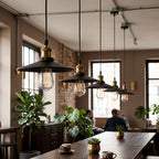 A row of industrial-style pendant lights with black metal conical shades, brass-toned hardware, and clear glass bulbs hanging over a wooden dining table in a modern loft cafe with people seated in the background.