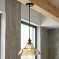 A modern industrial pendant light with a clear glass bell shade, warm exposed bulb, brass-colored socket, and black cord, hanging from a ceiling beam in a minimalist interior with concrete walls.