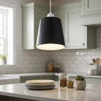 A black metal lamp shade hangs lit in a kitchen with white cabinets and a tiled backsplash.