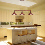 A modern kitchen island with a three-light linear pendant fixture hanging above. The pendant lights have a reddish-brown metal frame and three conical shades, illuminating a light wood kitchen island and two tan bar stools.