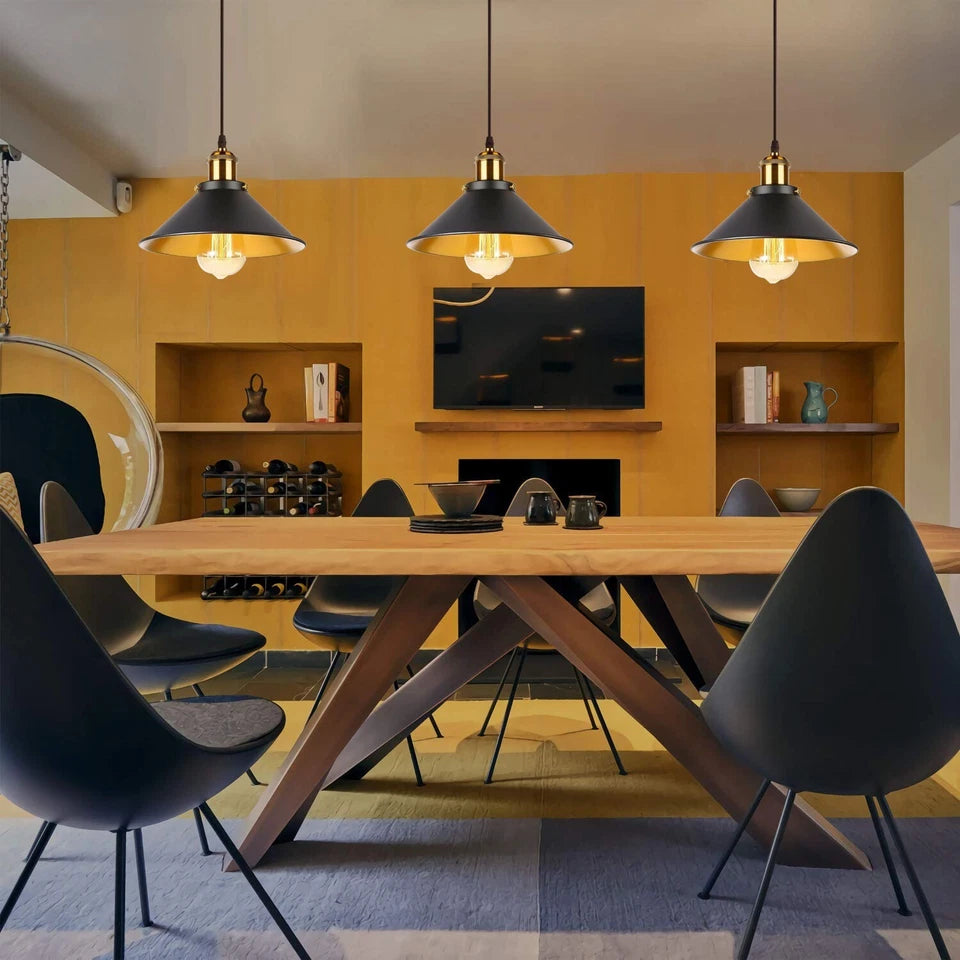 A modern dining room scene with a wooden dining table, black molded chairs, three black pendant lights with brass accents, and a warm yellow-orange accent wall behind a fireplace and TV.